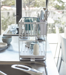 Side view of white Two-Tier Dish Rack on countertop holding dishware and cleaning accessories by Yamazaki Home. view 7