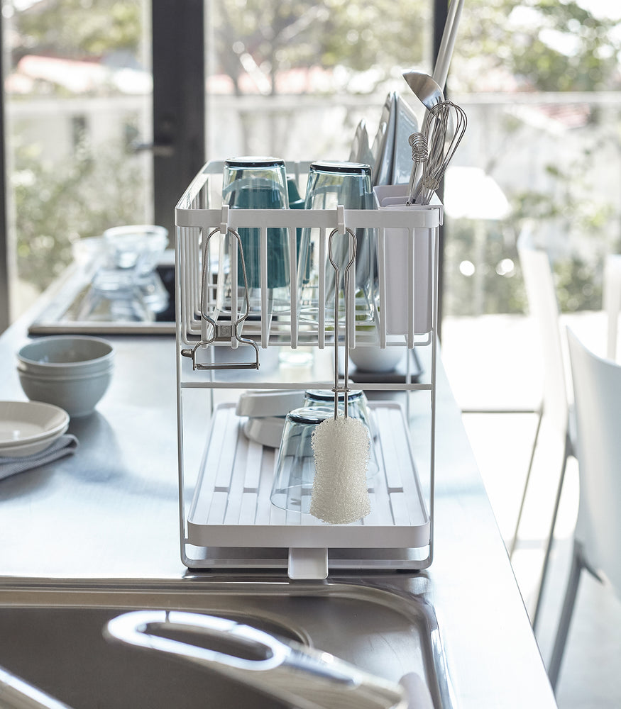 View 7 - Side view of white Two-Tier Dish Rack on countertop holding dishware and cleaning accessories by Yamazaki Home.