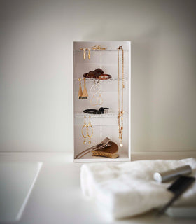 Sitting on top of a white bathroom counter is a jewelry organizer. It is a white resin rectangle with an open face and top featuring three transparent shelves distributed evenly with upward facing hooks along the edges, the last section of the rectangle is bare for added storage and has a small upward facing lip.. view 3