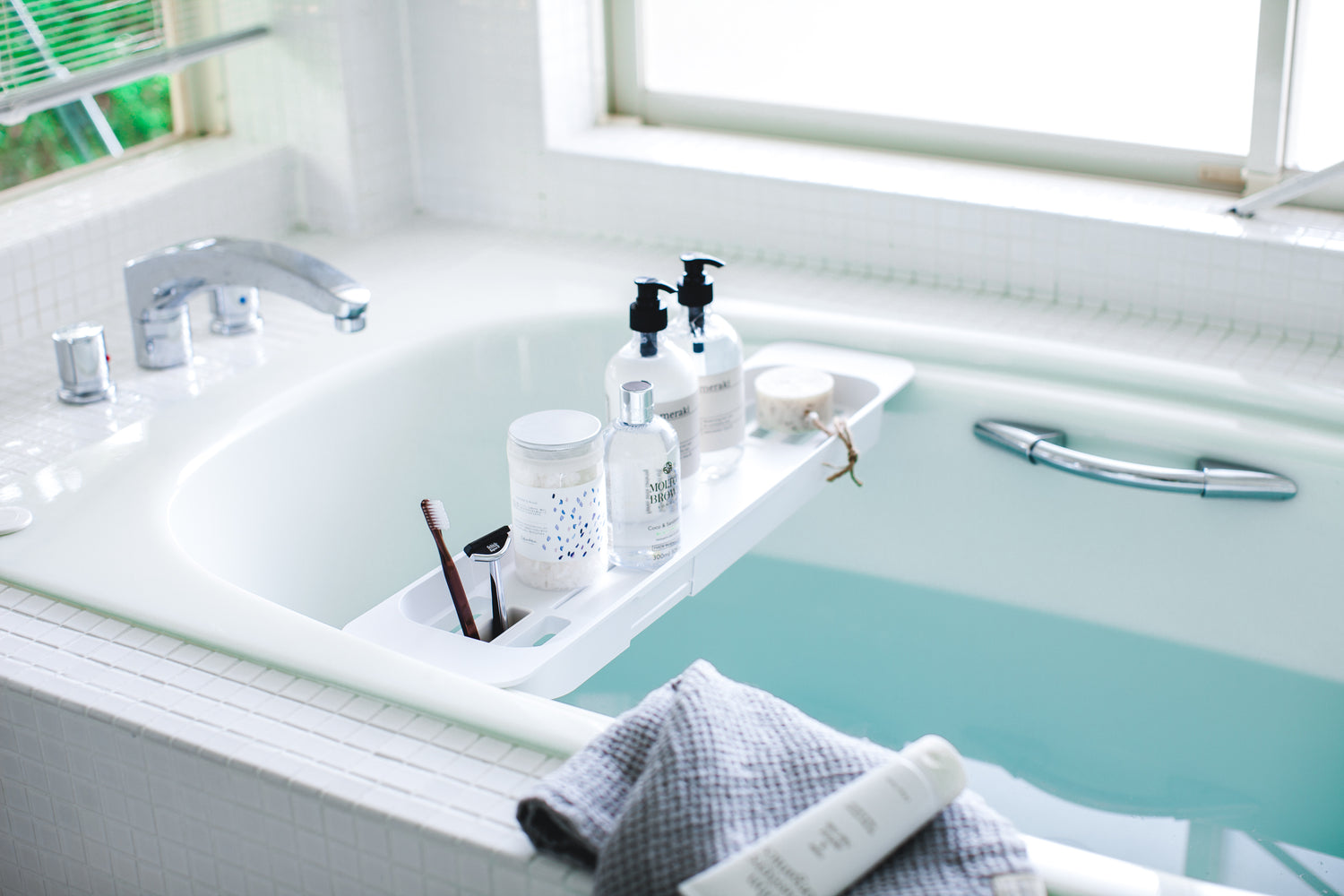 View 3 - White Expandable Bathtub Caddy holding beauty cleaning products in bathtub by Yamazaki Home.