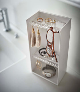 Looking down on top of a white bathroom counter is a white resin rectangular jewelry holder with an open face and top with two removable transparent shelves with upward facing hooks along the edge. The bottom has a small upward facing lip. The top shelf is holding a set of gold rings, on the hooks, a pair of earrings and a singular pair of reading glasses. The next shelf houses a black cord bracelet on the surface and on the hooks a pair of gold dangly earrings. The bottom houses a wristwatch. view 4