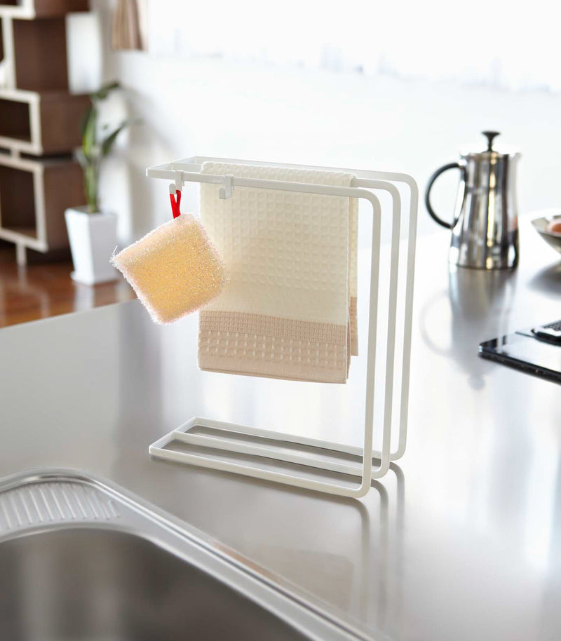 Front view of white Dish Towel Hanger holding towel and sponge on countertop by Yamazaki Home.