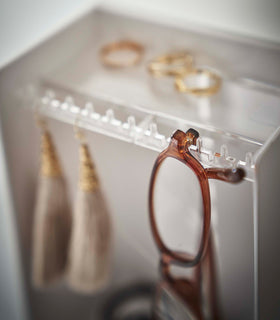 A detailed shot of the corner of a white plastic jewelry organizer. A transparent shelf with an upward facing lip is shown with hooks along the edge. Out-of-focus are three gold rings sitting on the shelf, and hanging on the hooks are a pair of out-of-focus earrings, in focus are the corner of a pair of reading glasses threaded along the hooks edge. view 5