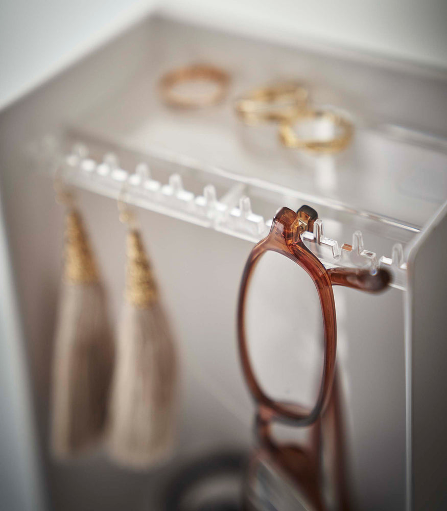 View 5 - A detailed shot of the corner of a white plastic jewelry organizer. A transparent shelf with an upward facing lip is shown with hooks along the edge. Out-of-focus are three gold rings sitting on the shelf, and hanging on the hooks are a pair of out-of-focus earrings, in focus are the corner of a pair of reading glasses threaded along the hooks edge.