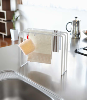 White Dish Towel Holder holding washcloths and sponge next to kitchen sink by Yamazaki Home. view 3