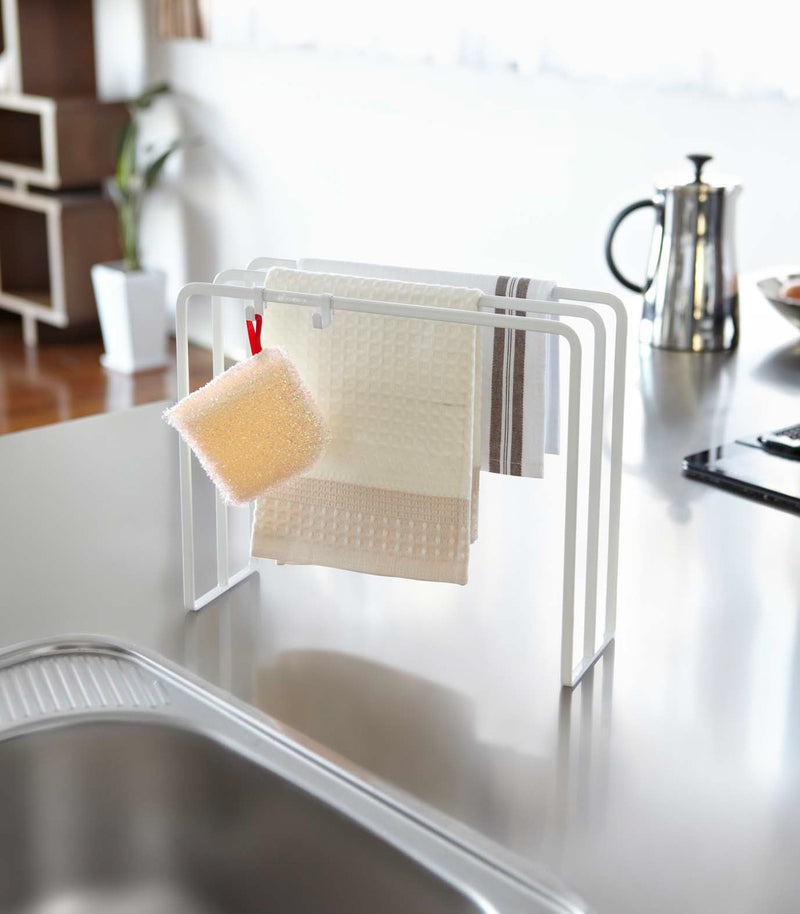 White Dish Towel Holder holding washcloths and sponge next to kitchen sink by Yamazaki Home.