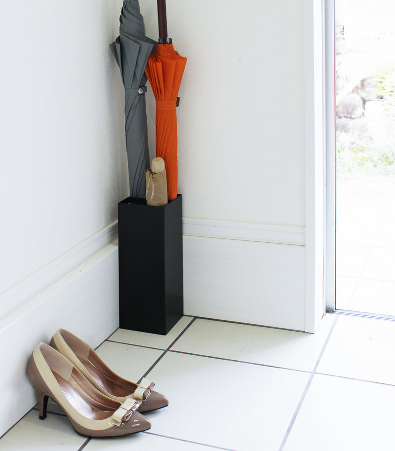 Compact Black Umbrella Stand in entryway holding two umbrellas, and one foldable umbrella by Yamazaki home.
