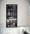 Sitting on top of a white bathroom counter is a black resin rectangle jewelry organizer. It has an open face and top with three transparent shelves with upward facing hooks along the edges, the last section of the rectangle is bare for added storage and has a small upward facing lip.