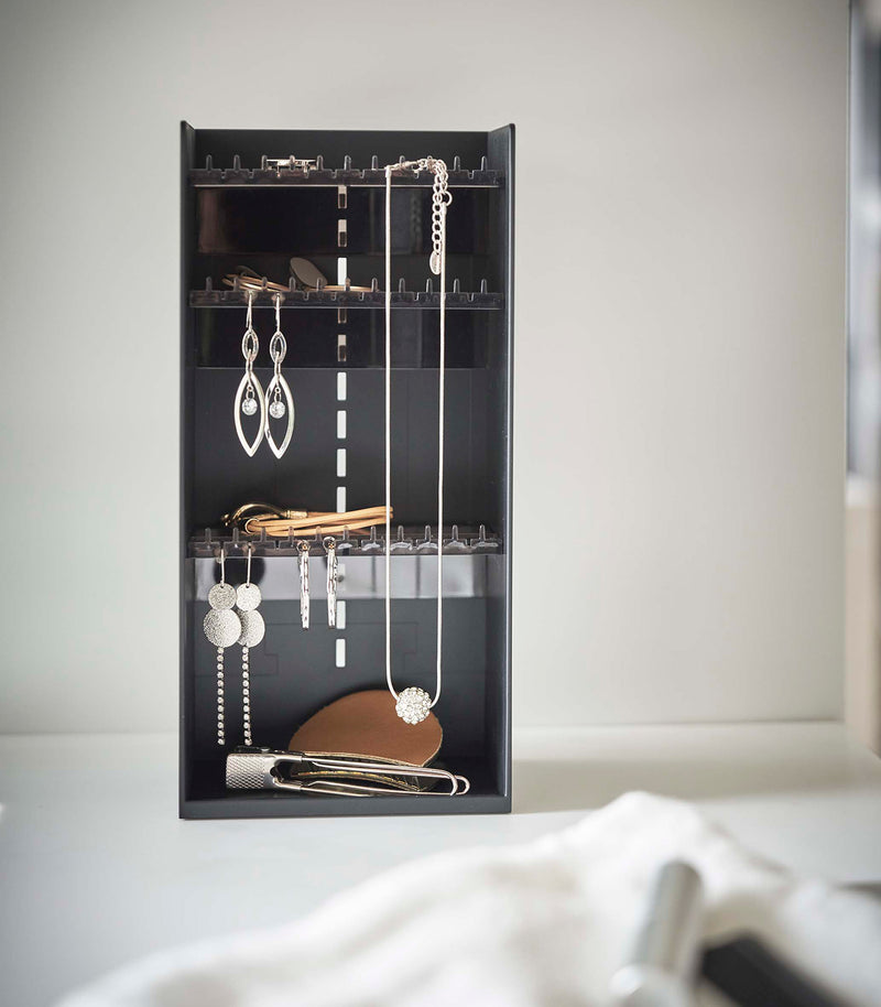 Sitting on top of a white bathroom counter is a black resin rectangle jewelry organizer. It has an open face and top with three transparent shelves with upward facing hooks along the edges, the last section of the rectangle is bare for added storage and has a small upward facing lip.
