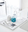 White Dish Drainer Tray holding silverware and cups on sink counter by Yamazaki Home.
