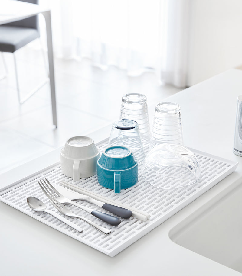 White Dish Drainer Tray holding silverware and cups on sink counter by Yamazaki Home.
