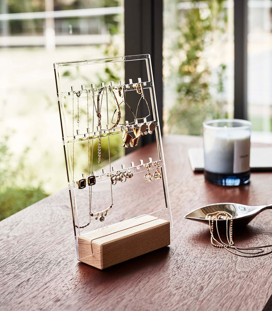 View 3 - A clear acrylic translucent earring holder with a rectangular light-colored wood base are displayed on a dark wood dresser. The acrylic holder has upward pointed hooks and slots placed in an interchangeable pattern. Hanging from the hooks are chained necklaces, and in the slots are various earrings. On the surface of the dresser, in front of the earring holder, is a leaf-shaped decorative catch-all plate with necklaces strewn inside.