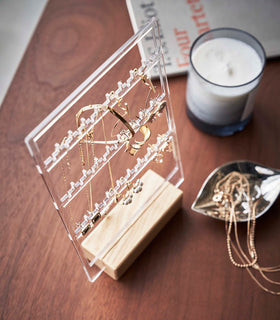 A birds-eye view of a clear acrylic translucent earring holder with a rectangular light-colored wood base on a dark wood dresser. The acrylic holder has upward pointed hooks and slots placed in an interchangeable pattern. Hanging from the hooks are chained necklaces, and in the slots are various earrings. On the surface of the dresser, in front of the earring holder, is a leaf-shaped decorative catch-all plate with necklaces strewn inside. view 4