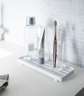 White slotted tray holding toothbrush and mouthcare items on bathroom sink counter by Yamazaki Home. view 2