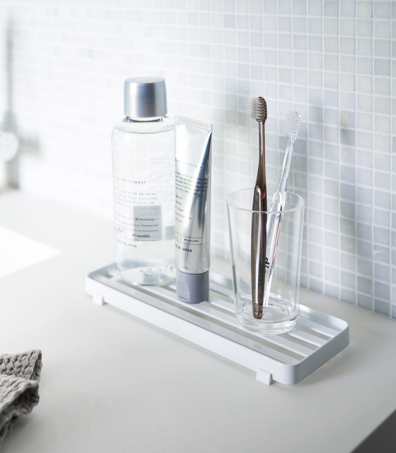 White slotted tray holding toothbrush and mouthcare items on bathroom sink counter by Yamazaki Home.