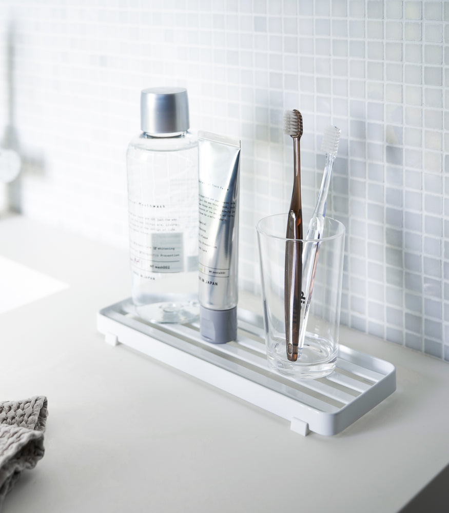 View 2 - White slotted tray holding toothbrush and mouthcare items on bathroom sink counter by Yamazaki Home.