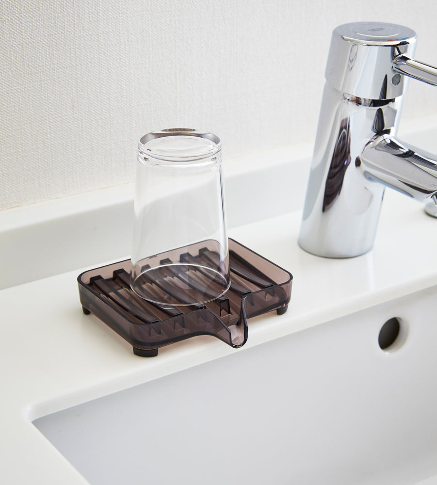 View 7 - Black Self-Draining Soap Tray holding glass on sink counter by Yamazaki Home.