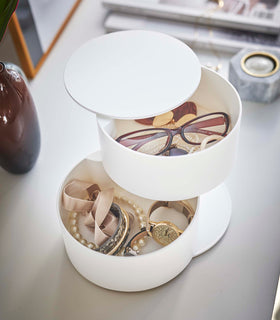 An angled bird’s-eye view of a two-tier white swivel accessory holder placed on a white dresser with a decorative vases and other décor seen in the background. The accessory holder’s tiers and lid are swiveled opened so the inside contents can be seen. In the first tier are a pair of reading glasses, earrings, and other various jewelry. The second tier holds a watch and an assortment of bracelets. view 5