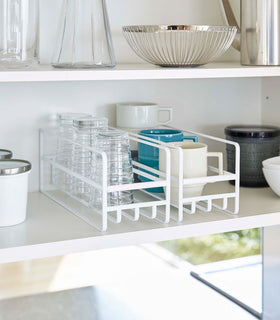 2 Yamazaki Home white Glass and Mug Cabinet Organizers on a shelf beside plates view 2