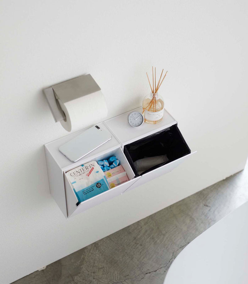 Aerial view of two open white Wall-Mount Storage bins holding items and trash bag in bathroom by Yamazaki Home.