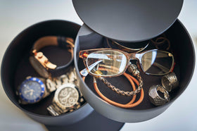 A detailed shot of the inside contents of a black two-tier swivel accessory holder. The tiers and lid are swiveled open so the inside contents can be seen. The focus of the image is the first-tier contents, there is a pair of reading glasses, two silver statement rings, and an assortment of corded bracelets. Out-of-focus in the second tier are three watches of different styles. Both compartments are lined with a felt liner. view 14