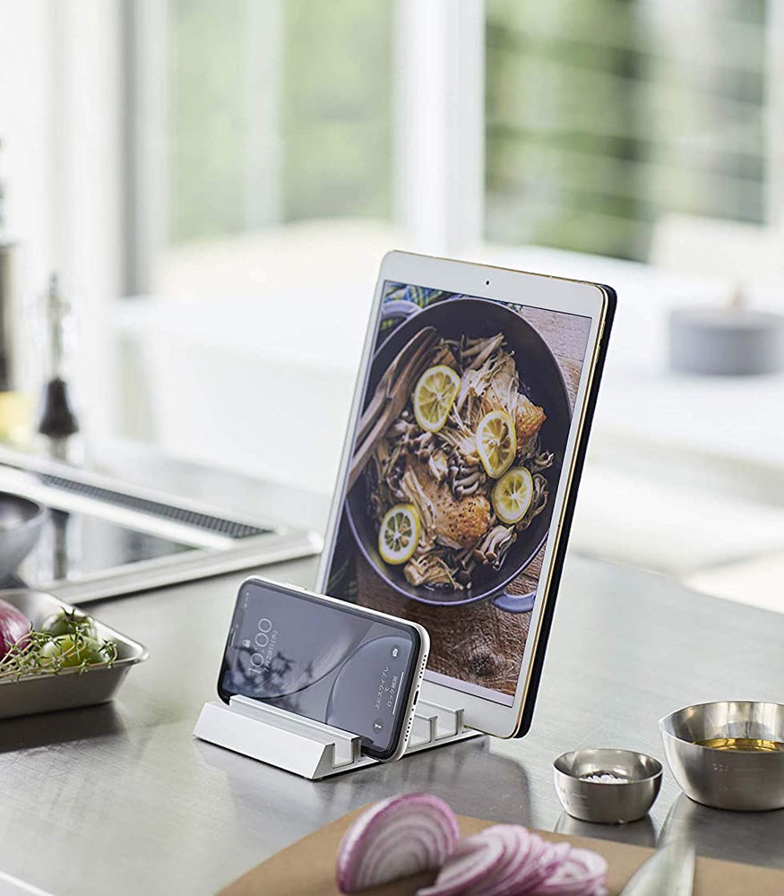 View 2 - White Phone and Tablet Stand holding tablet and phone on kitchen countertop by Yamazaki Home.