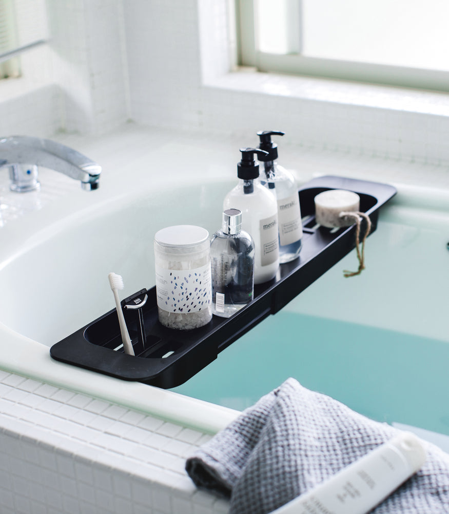View 9 - Black Expandable Bathtub Caddy holding bath products in bathroom by Yamazaki Home.
