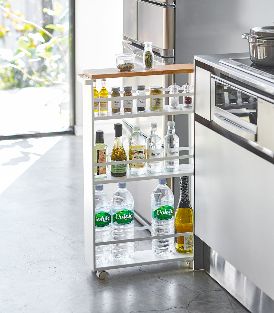 View 2 - Front view of white Rolling Storage Cart holding spices and liquid items in kitchen by Yamazaki Home.