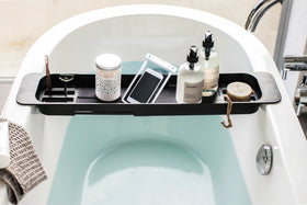 Front view of black Expandable Bathtub Caddy holding phone and cleaning products in bathroom by Yamazaki Home. view 10