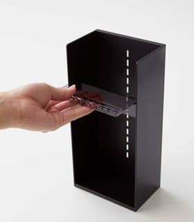 On a white surface is a black resin rectangular jewelry holder with an open face and top with a removable transparent shelf with upward facing along the edge. The bottom of the organizer has a small upward facing lip. A male hand pulls a transparent tray to adjust the location within the jewelry organizer. view 17