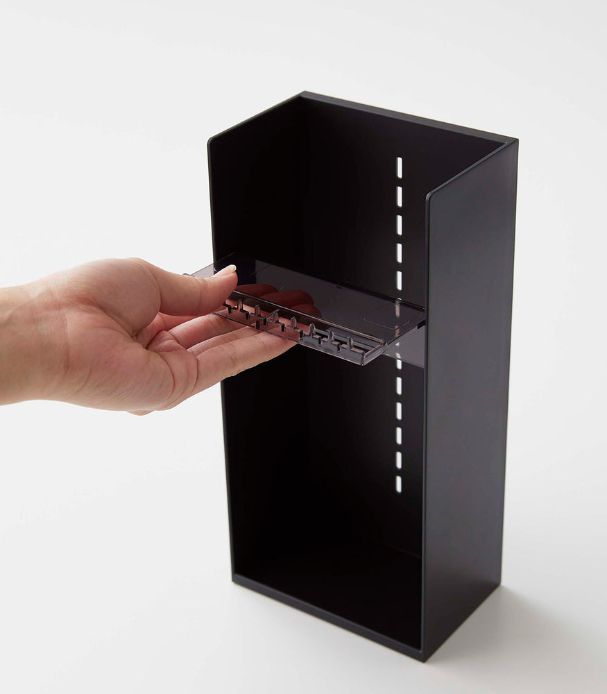 View 18 - On a white surface is a black resin rectangular jewelry holder with an open face and top with a removable transparent shelf with upward facing along the edge. The bottom of the organizer has a small upward facing lip. A male hand pulls a transparent tray to adjust the location within the jewelry organizer.